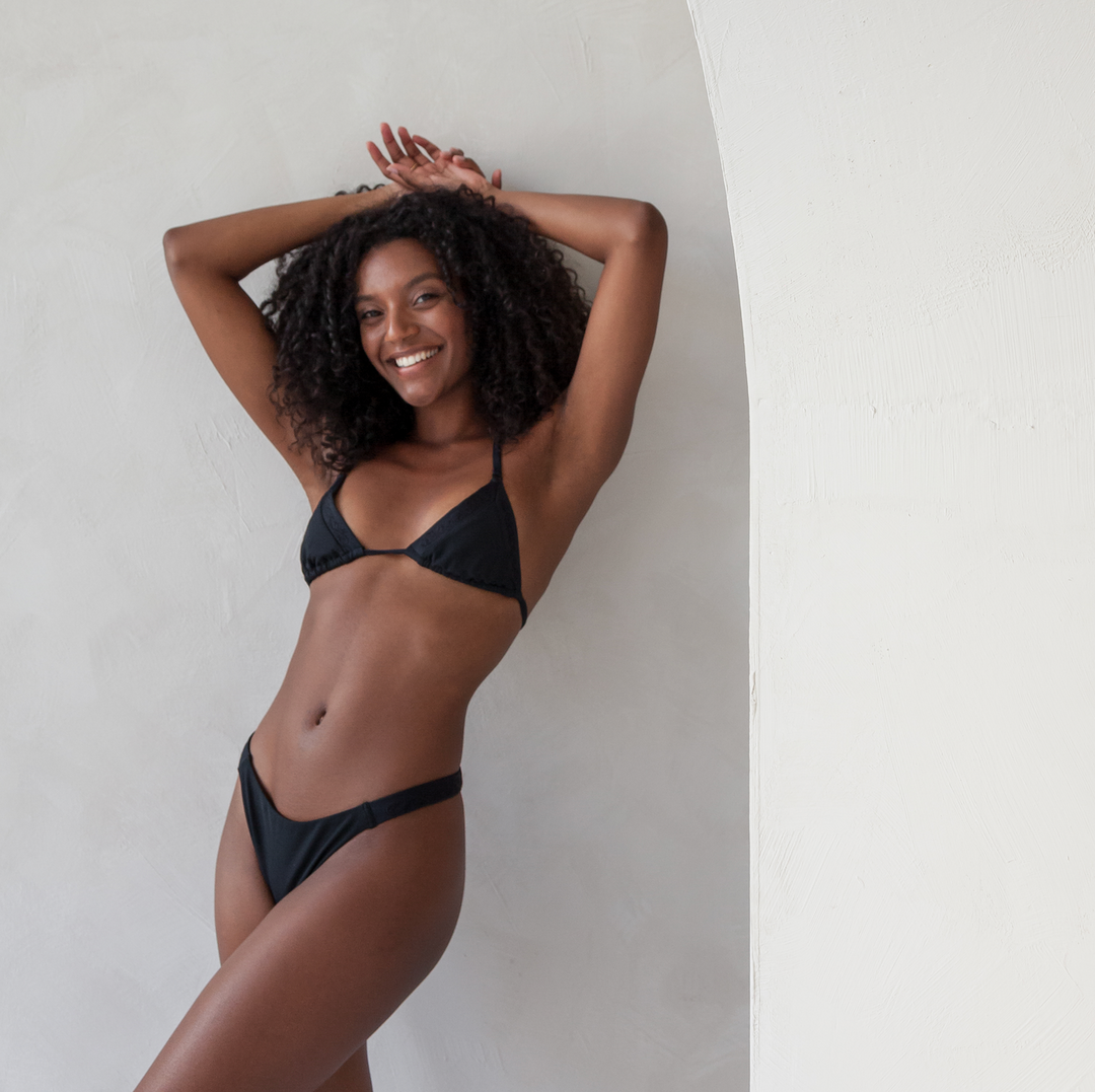 Woman wears the triangle bikini top with adjustable straps, and cheeky brazilian tanga bottom in black featuring rib fabric and satin stitch embroidery